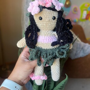 Pode incluir: Boneca de croch&ecirc; feita &agrave; m&atilde;o com uma coroa de flores e cabelo encaracolado preto. A boneca tem um rosto bronzeado, uma parte superior rosa e branca e uma saia verde. A boneca est&aacute; usando sand&aacute;lias rosa. A boneca tem aproximadamente 15 cm de altura.