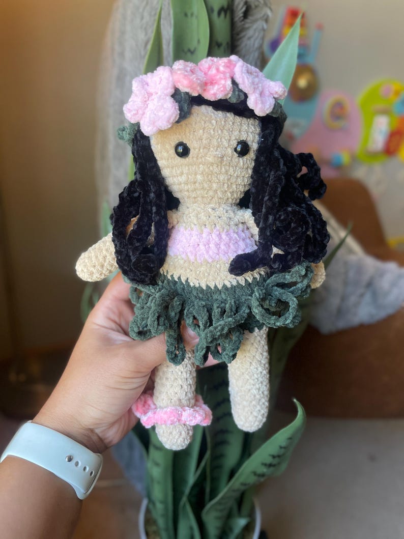 Pode incluir: Boneca de croch&ecirc; feita &agrave; m&atilde;o com uma coroa de flores e cabelo encaracolado preto. A boneca tem um rosto bronzeado, uma parte superior rosa e branca e uma saia verde. A boneca est&aacute; usando sand&aacute;lias rosa. A boneca tem aproximadamente 15 cm de altura.