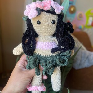 Pode incluir: Boneca de croch&ecirc; feita &agrave; m&atilde;o com corpo bege, cabelo preto e uma roupa rosa e verde. A boneca tem uma coroa de flores rosa e segura uma folha verde. A boneca tem aproximadamente 20 cm de altura.