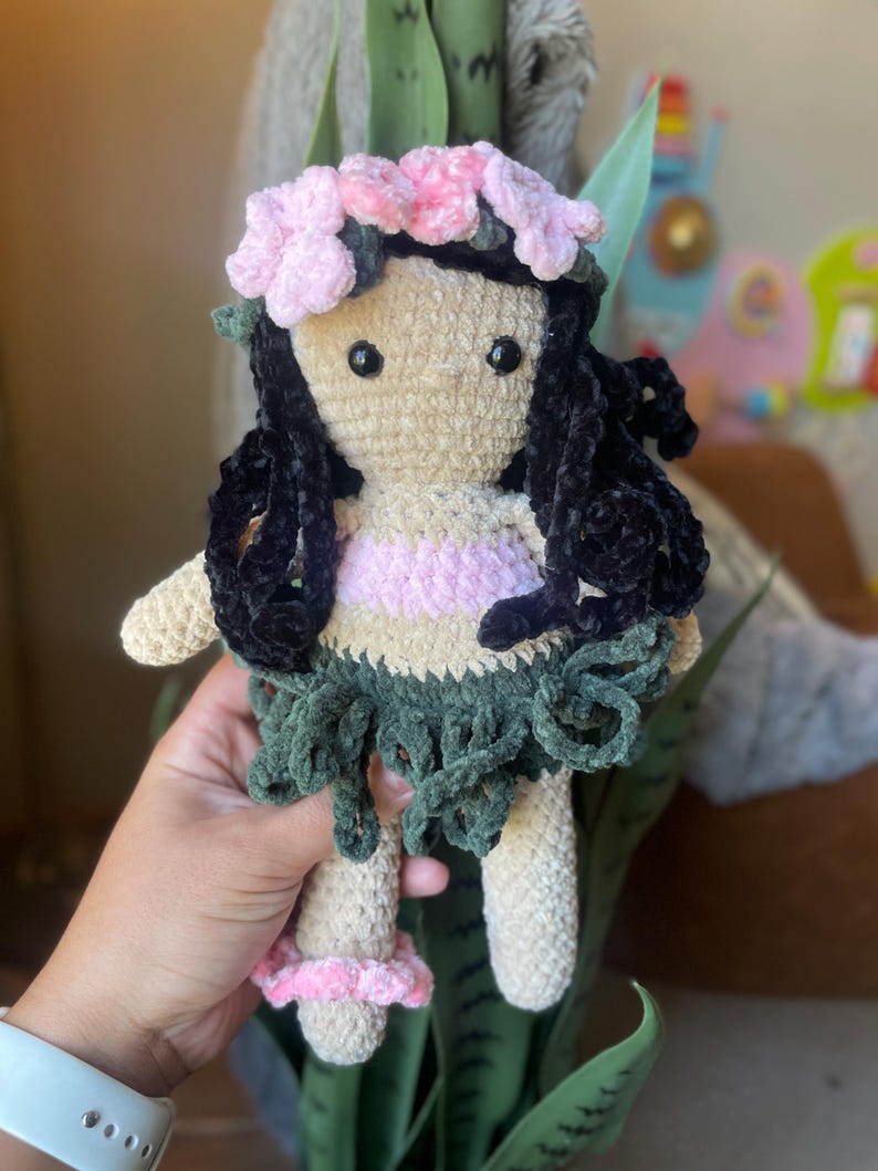 Pode incluir: Boneca de croch&ecirc; feita &agrave; m&atilde;o com corpo bege, cabelo preto e uma roupa rosa e verde. A boneca tem uma coroa de flores rosa e segura uma folha verde. A boneca tem aproximadamente 20 cm de altura.