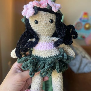 Pode incluir: Boneca de croch&ecirc; feita &agrave; m&atilde;o com corpo bege, cabelo preto e uma roupa rosa e verde. A boneca tem um adorno floral na cabe&ccedil;a e uma saia. A boneca est&aacute; sendo segurada por uma pessoa.
