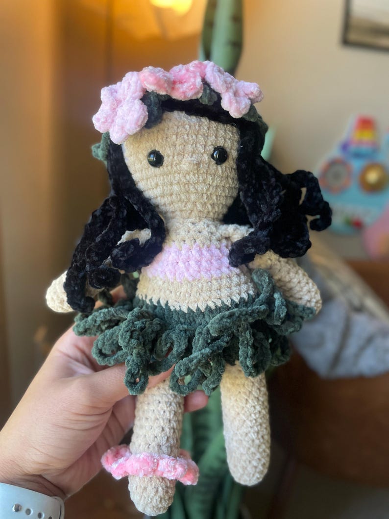 Pode incluir: Boneca de croch&ecirc; feita &agrave; m&atilde;o com corpo bege, cabelo preto e uma roupa rosa e verde. A boneca tem um adorno floral na cabe&ccedil;a e uma saia. A boneca est&aacute; sendo segurada por uma pessoa.