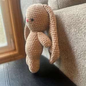 May include: A crocheted bunny toy in a light brown shade. The bunny features long, drooping ears, small arms, and a pink nose. The soft toy is placed on a black surface.