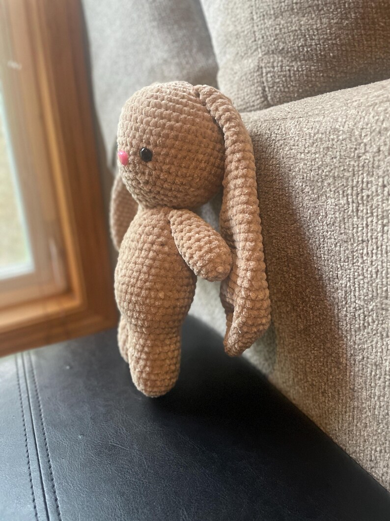 May include: A crocheted bunny toy in a light brown shade. The bunny features long, drooping ears, small arms, and a pink nose. The soft toy is placed on a black surface.