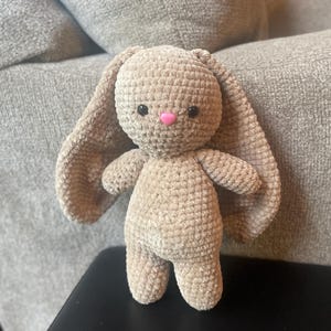 May include: A handmade, beige crochet bunny with long, floppy ears and a pink nose. The plush toy has black eyes and is standing upright. The bunny is made of a soft, textured yarn.