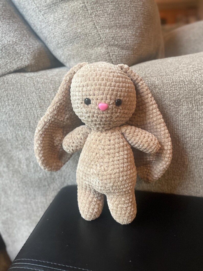 May include: A handmade, beige crochet bunny with long, floppy ears and a pink nose. The plush toy has black eyes and is standing upright. The bunny is made of a soft, textured yarn.