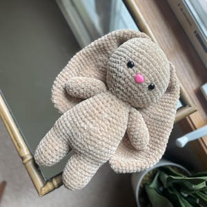 May include: A handmade, beige crochet bunny plush toy with long, floppy ears. The bunny has black safety eyes and a pink nose. The plush is lying on a mirror, with a window and a plant in the background.