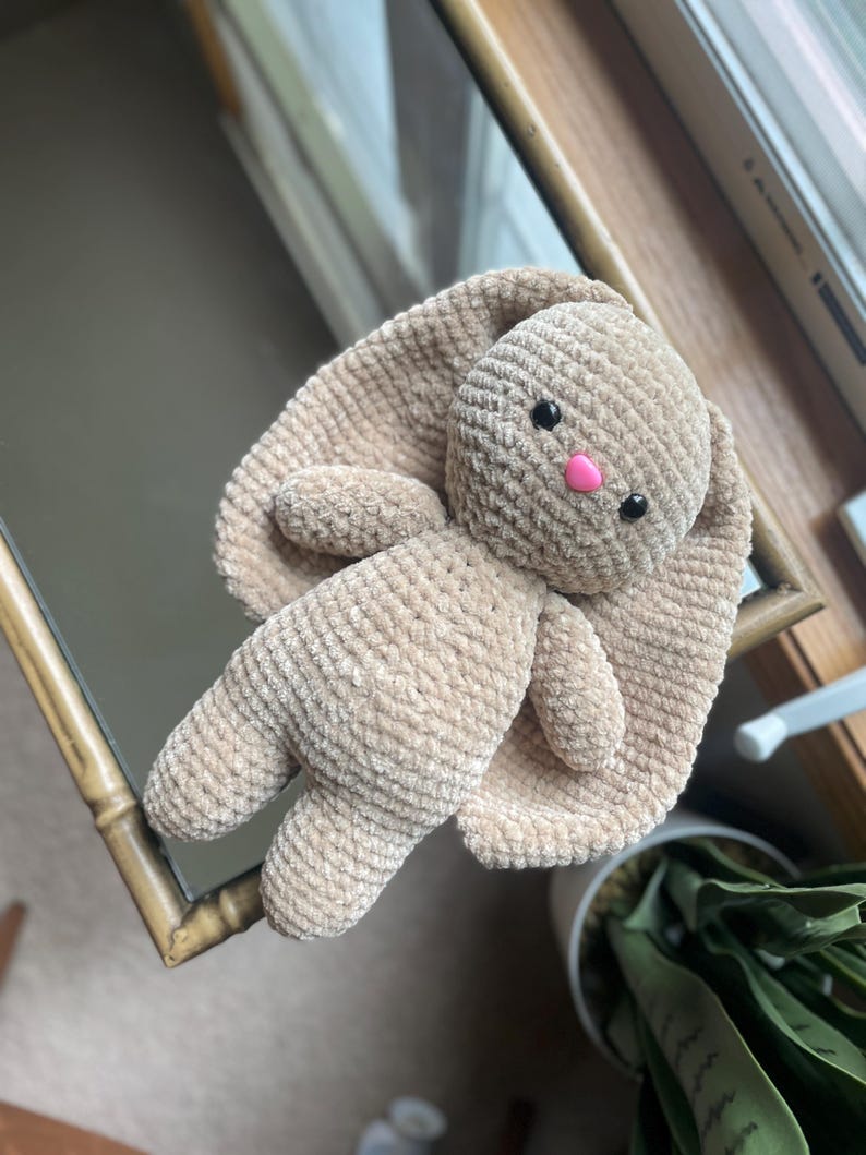 May include: A handmade, beige crochet bunny plush toy with long, floppy ears. The bunny has black safety eyes and a pink nose. The plush is lying on a mirror, with a window and a plant in the background.
