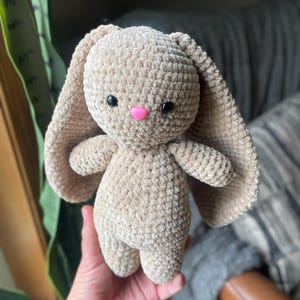May include: A beige crochet bunny with long, floppy ears, small arms, and legs. The bunny has black eyes and a pink nose. The plush toy is soft and cuddly, ideal for a child's toy collection.