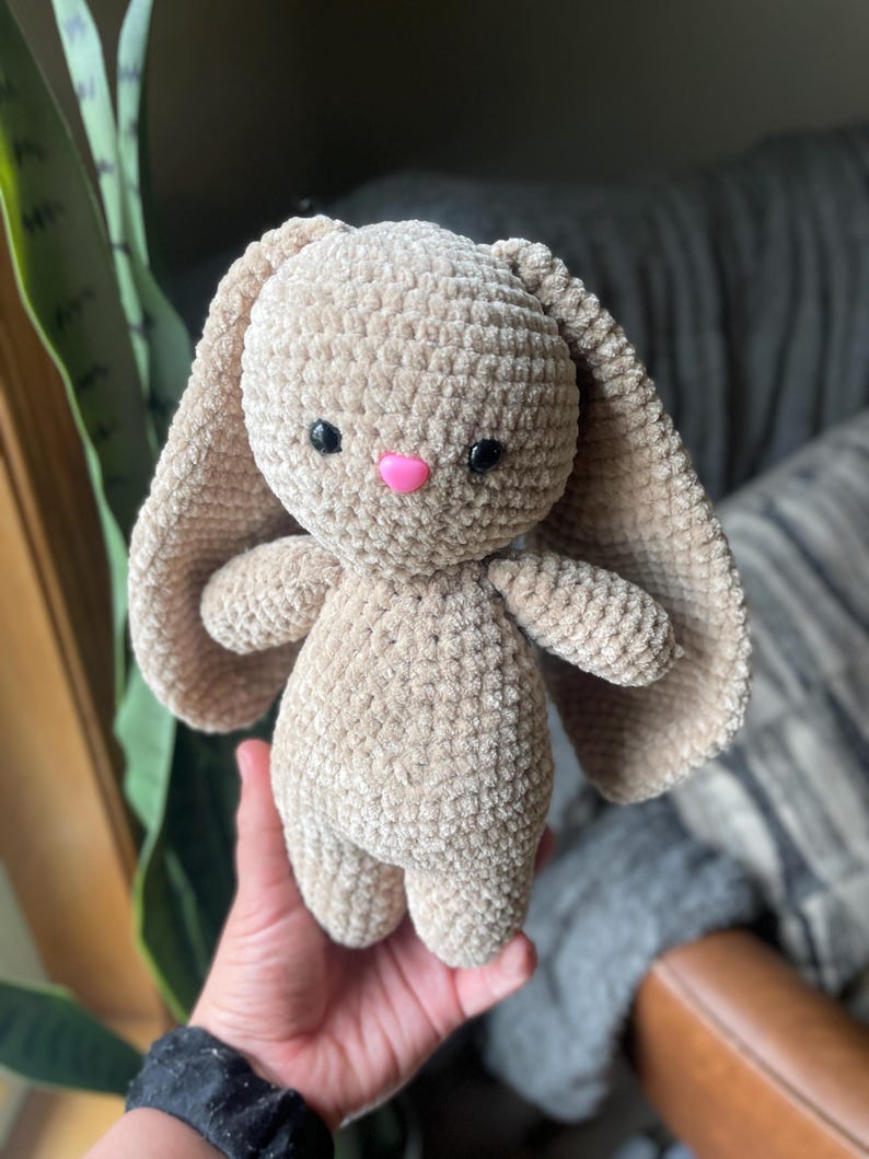 May include: A beige crochet bunny with long, floppy ears, small arms, and legs. The bunny has black eyes and a pink nose. The plush toy is soft and cuddly, ideal for a child's toy collection.