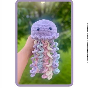 Op de afbeelding: Een gehaakt kwal speelgoed in tinten lavendel, roze, blauw en wit. De kwal heeft een afgeronde kop met twee kleine zwarte ogen en krullende tentakels. De tekst "JELLYFISH PATTERN" staat bovenaan.