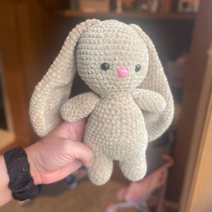 May include: A handmade, beige crochet bunny with long floppy ears, black eyes, and a pink nose. The plush toy is held up in front of a blurred background. The bunny is soft and cuddly, perfect for a child.