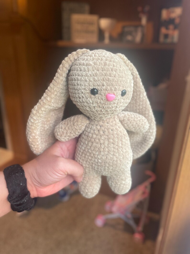 May include: A handmade, beige crochet bunny with long floppy ears, black eyes, and a pink nose. The plush toy is held up in front of a blurred background. The bunny is soft and cuddly, perfect for a child.