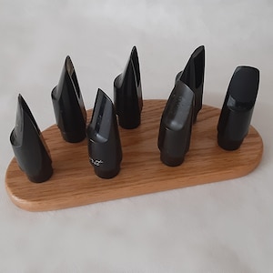 May include: A collection of black saxophone mouthpieces arranged on a light brown, oval-shaped wooden holder. The mouthpieces are glossy and have a conical shape. The holder is made of wood and has a smooth finish.