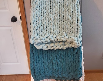 May include: Three chunky knit blankets stacked on a wooden ladder. The top blanket is light blue, the middle blanket is teal, and the bottom blanket is a pale pink.