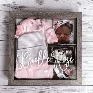 May include: A shadow box with baby items, including a pink onesie, a striped hat, and a photo of a newborn. The name "Brielle Rose" is displayed in white script. A pacifier and ultrasound image are also visible.