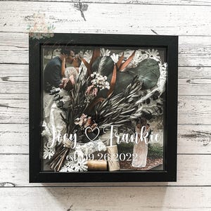 May include: A black shadow box frame with a wedding theme. The frame contains a photo of a couple, dried flowers, lace, and two corks. The names "Joey" and "Frankie" are written in cursive, along with the date "09.26.2022".