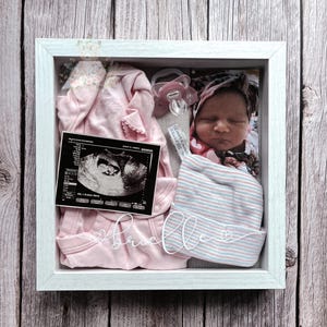 Könnte beinhalten: Ein weißer Schattenfugenrahmen mit einem rosa Baby-Body, einem rosa Schnuller, einem Neugeborenenfoto, einem Ultraschallbild und einer gestreiften Babymütze. Der Name Brielle steht in weißer Schreibschrift auf der Glasfront.