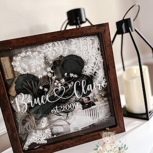 May include: A wooden shadow box with a glass front, containing a photo, lace, greenery, and a pearl necklace. The box is personalized with the names "Bruce & Claire" and the date "est. 2009".