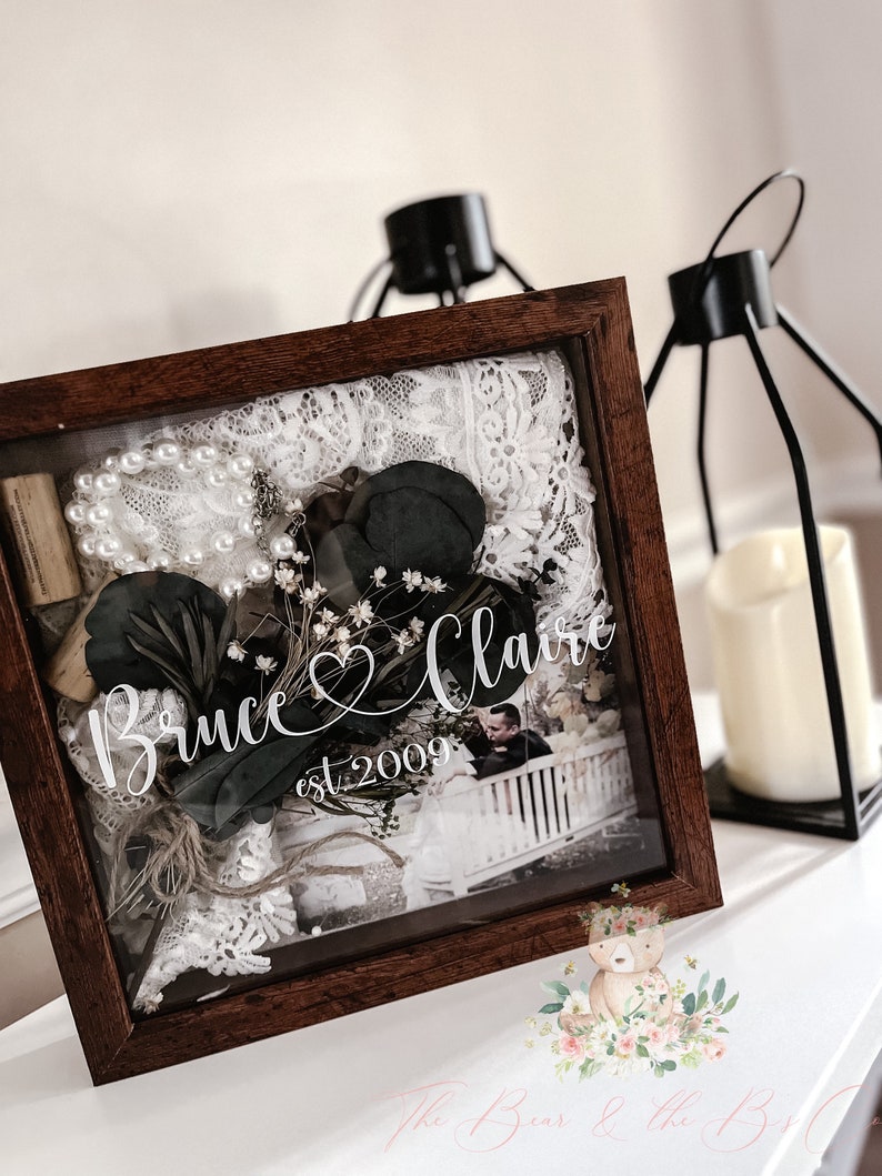 May include: A wooden shadow box with a glass front, containing a photo, lace, greenery, and a pearl necklace. The box is personalized with the names "Bruce & Claire" and the date "est. 2009".
