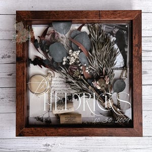 May include: A shadow box with a dark wood frame, showcasing a dried flower bouquet, eucalyptus leaves, and a pocket watch. The display includes a wedding invitation and wine corks. The text "HEDRICKS" and "10.30.2009" are visible.