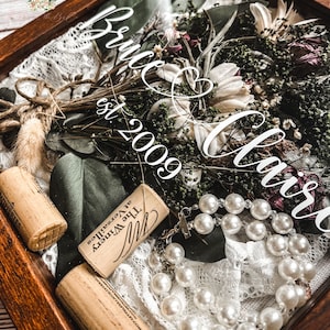 May include: A wooden shadow box with a dried flower bouquet, two wine corks, and a pearl necklace. The text "Bruce & Christ est. 2009" is visible on the glass.