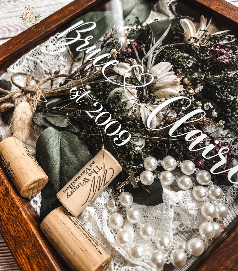May include: A wooden shadow box with a dried flower bouquet, two wine corks, and a pearl necklace. The text "Bruce & Christ est. 2009" is visible on the glass.