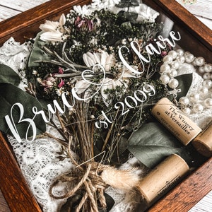 May include: A wooden shadow box with a dried flower bouquet, white lace, a string of pearls, and wine corks. The box has a white inscription that reads "Bruce & Claire est 2009".