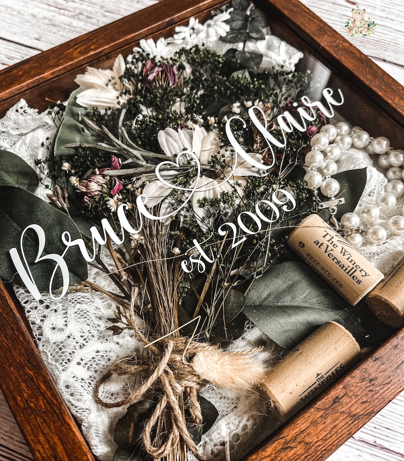 May include: A wooden shadow box with a dried flower bouquet, white lace, a string of pearls, and wine corks. The box has a white inscription that reads "Bruce & Claire est 2009".