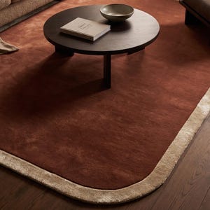 May include: A large, rectangular area rug with a deep terracotta-colored center and a wide, plush beige border. The rug features rounded corners and a soft, textured pile. A dark brown circular coffee table sits on the rug with a book and bowl.