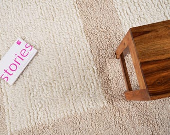 Pode incluir: Um tapete texturizado, branco sujo e bege, com um design de listras verticais. Uma pequena mesa quadrada de madeira está no canto superior direito. Um livro branco com a palavra rosa "stories" está no canto inferior esquerdo.