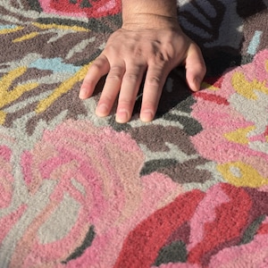Close-up photograph of a hand gently pressing into the dense, plush pile of a hand-tufted wool floral rug to showcase texture and softness. Detailed rug edge showing the dense wool pile, finished binding and cotton-canvas backing