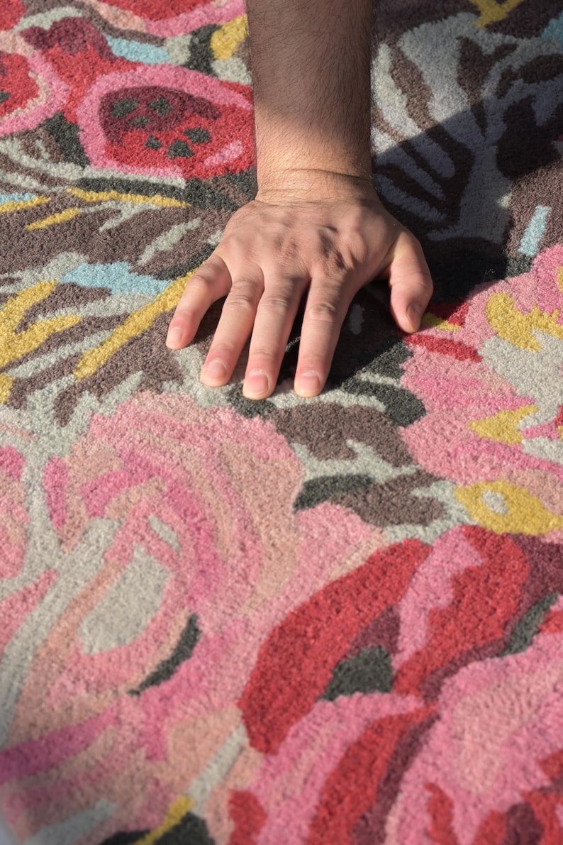 Close-up photograph of a hand gently pressing into the dense, plush pile of a hand-tufted wool floral rug to showcase texture and softness. Detailed rug edge showing the dense wool pile, finished binding and cotton-canvas backing