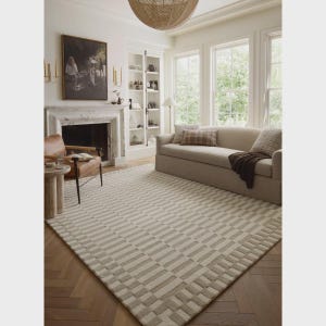 Könnte beinhalten: Ein beigefarbener Teppich mit geometrischem Muster dominiert den Raum. Das Design des Teppichs zeigt abwechselnd helle und mittelbeige rechteckige Blöcke. Der Raum umfasst auch ein beiges Sofa, einen Ledersessel, einen Kamin und ein großes Gemälde.