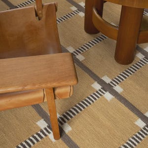 Könnte beinhalten: Ein gewebter Teppich in Beige, Braun und Weiß mit einem großflächigen Karomuster. Der Teppich liegt unter einem Holzstuhl mit beigem Ledersitz und einem runden Holztisch mit dicken zylindrischen Beinen und einer schraffierten Oberfläche.