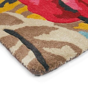 Macro photograph of the corner of a hand-tufted wool rug on a white studio background showing thick yarn fibers and cut-pile texture. Close-up of the hand tufted wool pile showing pink flowers and aqua botanical detailing.