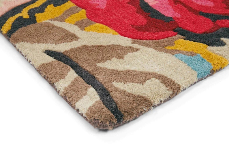 Macro photograph of the corner of a hand-tufted wool rug on a white studio background showing thick yarn fibers and cut-pile texture. Close-up of the hand tufted wool pile showing pink flowers and aqua botanical detailing.