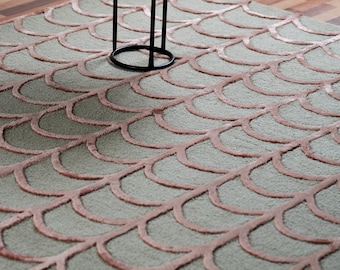 May include: A sage green area rug with a repeating, scalloped pattern in a muted rose color. The rug's design is reminiscent of fish scales. A black metal table leg is visible in the image.