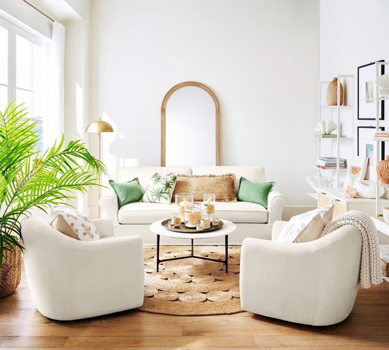 May include: A white living room with two white armchairs, a white sofa, a coffee table with candles, a large green plant, and a wooden arched mirror.