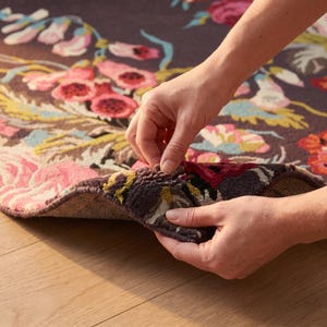 Close-up photograph of two hands lifting and bending the corner of a hand-tufted wool rug to demonstrate flexibility and pile thickness.