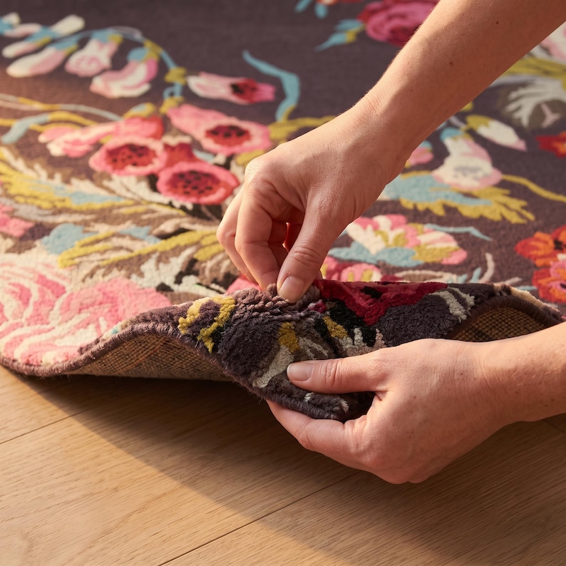 Close-up photograph of two hands lifting and bending the corner of a hand-tufted wool rug to demonstrate flexibility and pile thickness.