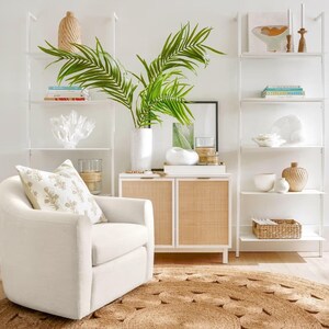 May include: A white swivel chair with a beige pillow sits in front of a white cabinet with woven rattan doors. The cabinet is next to a white ladder shelf with books, decorative items, and a potted palm plant.