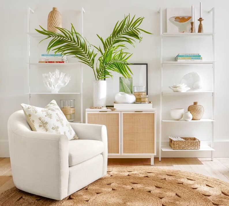 May include: A white swivel chair with a beige pillow sits in front of a white cabinet with woven rattan doors. The cabinet is next to a white ladder shelf with books, decorative items, and a potted palm plant.