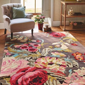 Editorial interior photograph of a floral hand-tufted area rug placed in a sunny living room beneath a modern lounge chair. Multicolor hand tufted floral rug made from New Zealand wool with a dark background and oversized pink flowers.