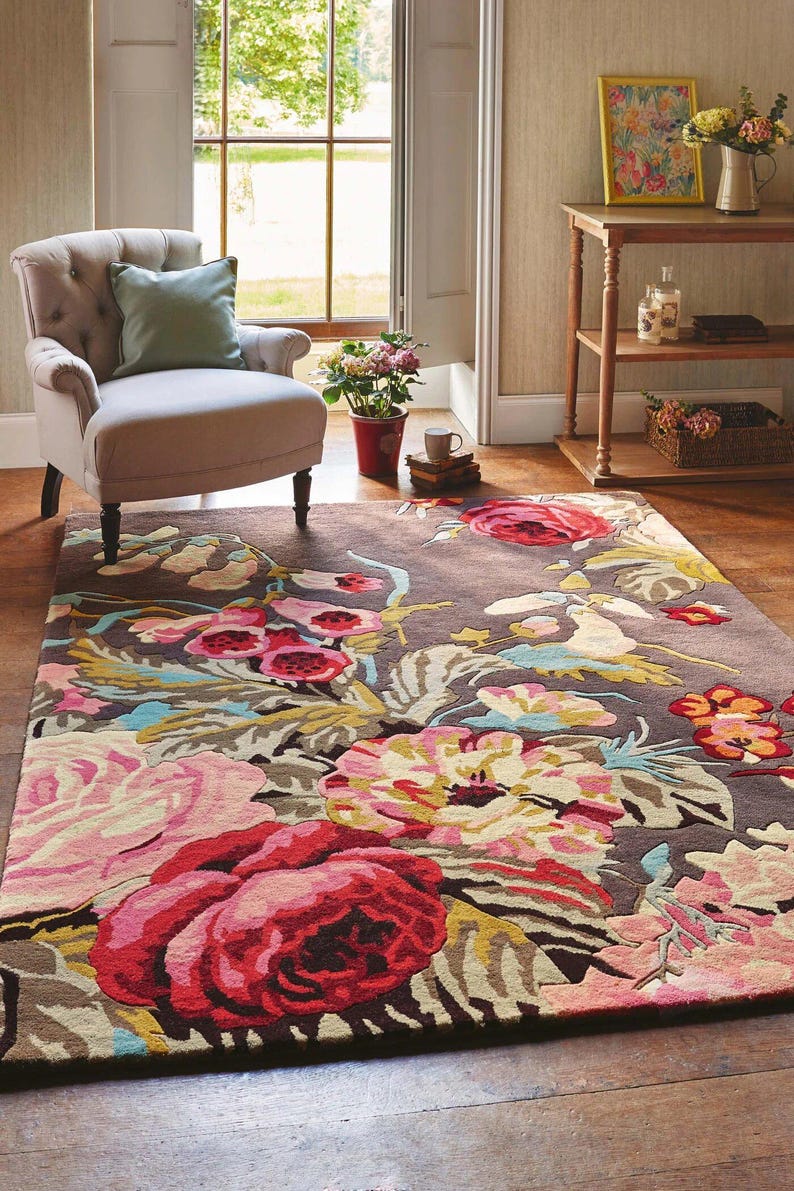 Editorial interior photograph of a floral hand-tufted area rug placed in a sunny living room beneath a modern lounge chair. Multicolor hand tufted floral rug made from New Zealand wool with a dark background and oversized pink flowers.