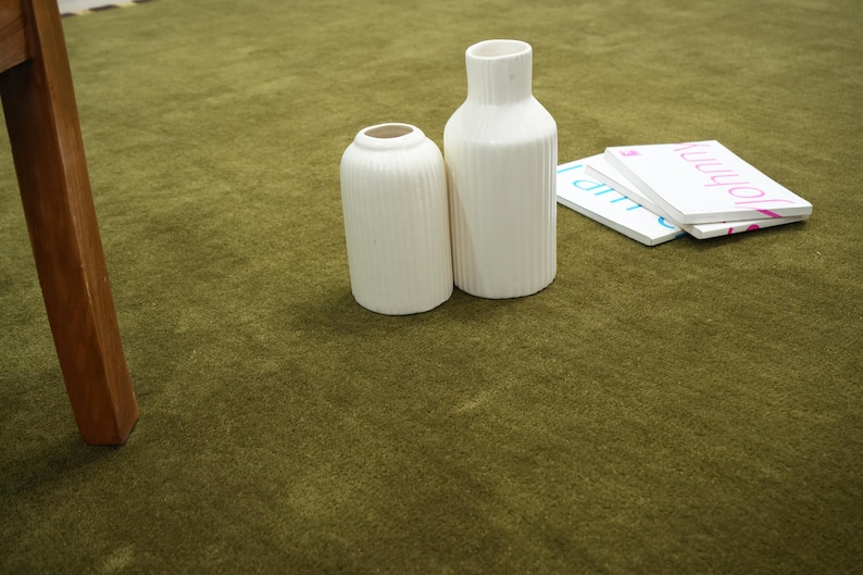 Peut inclure: Deux vases en c&eacute;ramique blanche avec des d&eacute;tails c&ocirc;tel&eacute;s verticaux sont pos&eacute;s sur une surface verte. &Agrave; c&ocirc;t&eacute;, de petits livres blancs avec des noms imprim&eacute;s en rose et bleu. Un pied de table en bois est visible.