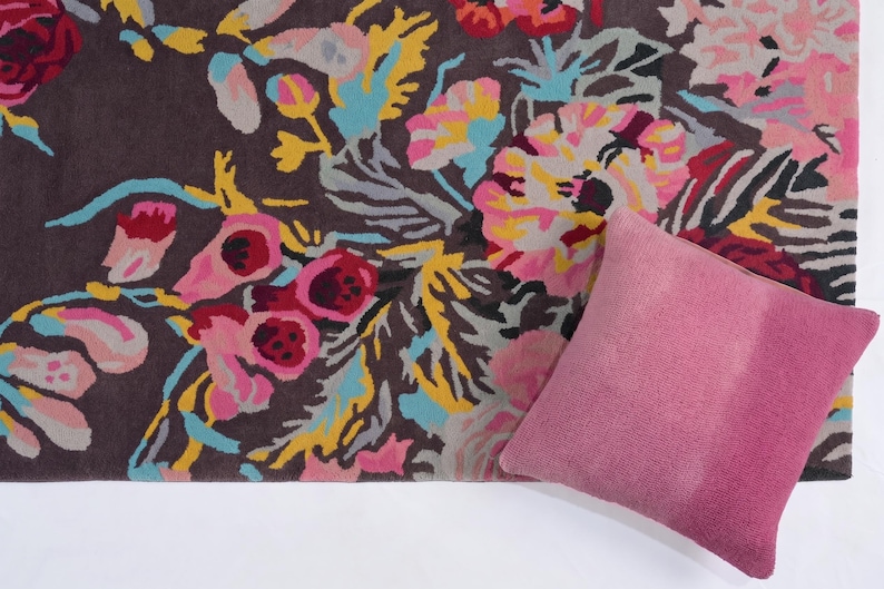 Partial top-down view of a floral hand-tufted rug styled on a light wooden floor with a dusty pink accent pillow placed on top. Summer Bloom multicolor floral rug displayed as a statement carpet for a living room or bedroom
