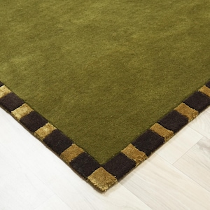 Peut inclure: Tapis vert olive avec une bordure marron fonc&eacute; et dor&eacute;e. Le tapis a une texture douce et un motif g&eacute;om&eacute;trique. Le coin du tapis est visible, montrant le motif d&eacute;taill&eacute; de la bordure.