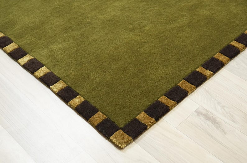 Peut inclure: Tapis vert olive avec une bordure marron fonc&eacute; et dor&eacute;e. Le tapis a une texture douce et un motif g&eacute;om&eacute;trique. Le coin du tapis est visible, montrant le motif d&eacute;taill&eacute; de la bordure.