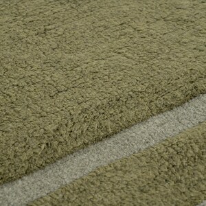 Pode incluir: Close-up de um tapete verde-oliva texturizado com uma faixa diagonal cinza claro. O tapete tem uma apar&ecirc;ncia macia e felpuda, sugerindo uma textura macia e confort&aacute;vel. O design &eacute; simples e moderno.